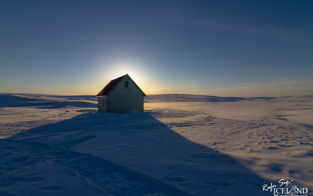 A small, pale green house with a red roof sits in a vast expanse of snowy landscape. The sun is setting in the background, casting a warm glow around the house. Shadows stretch across the snow, highlighting the undulating surface. The sky transitions from light blue to a deeper shade near the horizon, creating a tranquil winter scene.