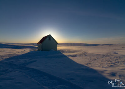 A small, pale green house with a red roof sits in a vast expanse of snowy landscape. The sun is setting in the background, casting a warm glow around the house. Shadows stretch across the snow, highlighting the undulating surface. The sky transitions from light blue to a deeper shade near the horizon, creating a tranquil winter scene.