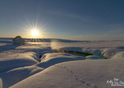 A serene winter landscape features a small, blue house in the distance, surrounded by a vast expanse of snow. The sun is setting, casting a warm, golden glow and creating starbursts in the sky. In the foreground, the snow is undulating, with a few visible animal tracks leading toward a crack in the snow, revealing a steaming hot spring nearby. The scene captures the tranquility and beauty of a cold, snowy environment.