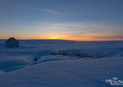 A serene winter landscape featuring a small, solitary house with a red roof, surrounded by a vast expanse of snow. In the background, the sun sets near the horizon, casting warm orange and yellow hues across the sky. The snow-covered ground has gentle undulations, and a small stream can be seen flowing nearby, with steam rising from its surface. The overall scene conveys a peaceful, isolated atmosphere in a wintry setting.