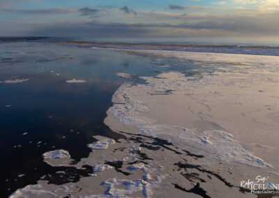 A tranquil scene of icy waters where patches of ice float on a reflective surface. The landscape features a mix of dark water and light ice, with the horizon gently fading into a soft, cloudy sky. The light suggests either dawn or dusk, casting a serene ambiance over the entire setting.