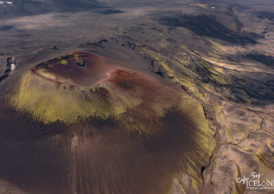 An aerial view of a volcanic landscape featuring a prominent conical volcano with a crater at the summit. The volcano exhibits a mix of dark volcanic rock and patches of green moss or vegetation, contrasting with the arid, black terrain surrounding it. The rugged landscape extends into the distance, with a textured surface that includes lava flows and valleys, under a clear sky.
