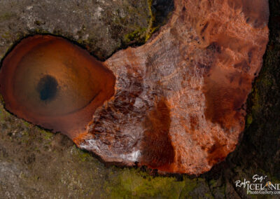 An aerial view of a geothermally heated area featuring a circular, deep body of water surrounded by reddish-brown mineral deposits. The edges of the water are contrasted by patches of dark, rocky terrain and green moss, highlighting the unique geological features of the landscape.