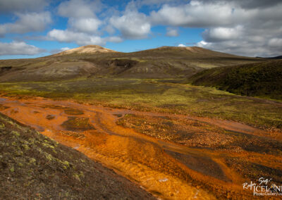 A vibrant landscape featuring rolling hills covered in a mix of green moss and brown earth. In the foreground, a winding river with orange hues flows through the scene, contrasting against the dark terrain. The sky is partly cloudy with a mix of blue and white clouds. In the background, a mountain rises gently, completing the striking natural scenery.