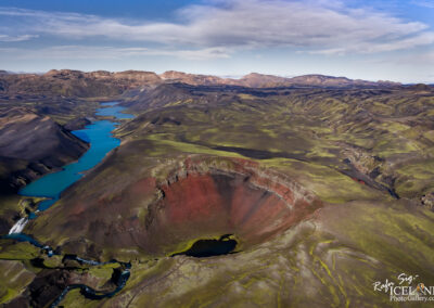 An aerial view of a dramatic landscape featuring a volcanic crater with reddish-brown walls, surrounded by lush green hills and a deep blue river winding through the valley. Mountains rise in the background under a partly cloudy sky, highlighting the natural beauty of the area. Small waterfalls can be seen cascading down the hillsides, creating a contrast with the surrounding terrain.