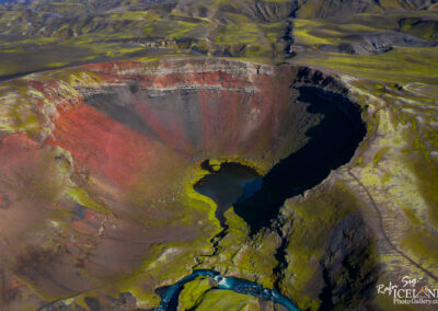 An aerial view of a volcanic crater featuring vibrant reds, blacks, and greens in the rocky landscape. The crater has a small lake at its base, surrounded by patches of moss and lichen, and a winding river flows along the foreground. The surrounding terrain is characterized by rolling hills and diverse geological formations.
