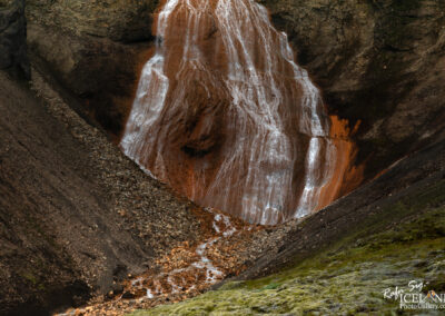 A cascading waterfall flows down a rocky cliff, with water trickling over reddish-brown layers of rock. The surrounding landscape features dark, steep terrain, dotted with pebbles and greenery at the base. The scene conveys a sense of natural beauty and rugged wilderness.