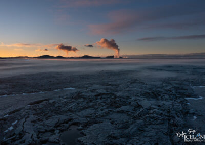 A dramatic landscape featuring a rugged, dark lava field in the foreground, with patches of frost and water. In the background, rolling hills and mountains are visible beneath a colorful sky at sunset. Clouds are illuminated by the setting sun, with steam rising from geothermal activity, adding a mystical touch to the scene. The overall atmosphere conveys a sense of tranquility and the raw power of nature.