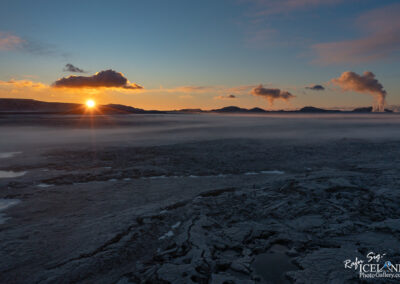 A stunning landscape featuring a sunset over a rugged terrain, characterized by textured dark volcanic rock and mist rising from the ground. In the distance, low hills are silhouetted against a colorful sky with orange and blue hues, and clouds scattered throughout. Two steam plumes can be seen rising from the ground, adding an otherworldly feel to the scene.