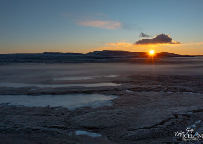 A serene landscape featuring a sunset over a vast, rocky terrain. The sun is just above the horizon, casting warm golden rays and illuminating a few clouds in the sky. The land appears desolate with patches of ice and mist, suggesting a cold environment, while the distant hills frame the scene, partially highlighted by the fading light.