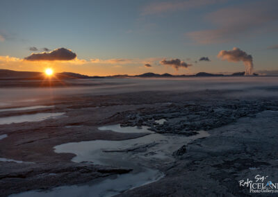 A breathtaking sunset over a rocky landscape with patches of water, set against a backdrop of distant mountains and clouds. The sun is partially hidden behind the horizon, casting a warm, golden glow across the scene, while steam rises from a geothermal area in the distance, adding an ethereal quality to the atmosphere. The foreground features textured rock formations and reflective pools that enhance the rugged beauty of the terrain.