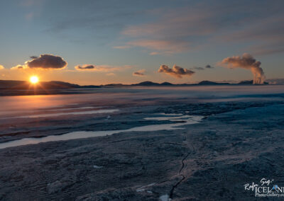 A serene landscape at sunset featuring a glowing sun on the horizon, casting warm light over a rugged terrain. The foreground shows textured ground with patches of water and dried earth, while distant mountains rise under a partially cloudy sky. Smoke billows from a geothermal area on the right, adding to the natural beauty of the scene.