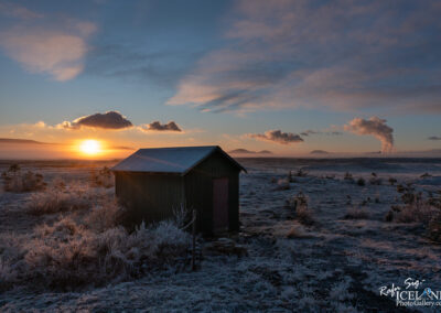 A small green shed stands in a frost-covered field with a sunrise in the background. The sun casts a warm orange glow across the horizon, while clouds float in the sky. In the distance, low hills and steam rising from geothermal activity are visible, creating a serene and picturesque landscape.