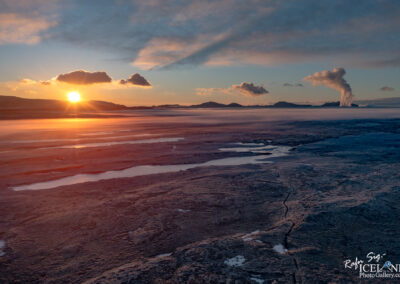 A breathtaking landscape at sunset, featuring a vibrant sun hovering above distant mountains. The foreground showcases a rugged, textured terrain with patches of gleaming water, while a plume of steam rises from a geothermal area in the background. Soft clouds are scattered across the sky, reflecting the warm hues of the setting sun, creating a serene and otherworldly atmosphere.
