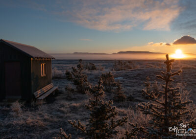 A small green wooden cabin sits in a frosty landscape, with a wooden bench in front. In the background, soft hills are partially shrouded in mist, and the sun is rising above the horizon, casting warm golden light across the scene. Patchy frost covers the ground and surrounding small evergreen trees, while the sky displays a gradient of colors from blue to warm orange and yellow near the sun.
