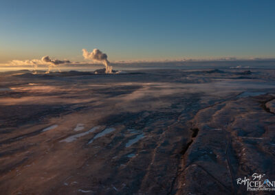 Aerial view of a vast, barren landscape with subtle textures and colors, illuminated by the warm light of sunset. The scene features several plumes of steam rising from geothermal activity in the distance, contrasting against the rocky terrain. Areas of water catch the light, while distant mountains and a horizon line complete the picturesque yet rugged setting.