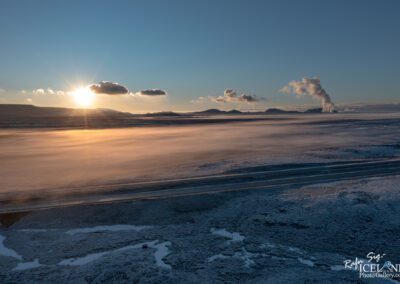 A panoramic view of a scenic landscape at sunset, featuring a vast expanse of frozen terrain with patches of snow and water. In the background, rolling hills and mountains are silhouetted against the sky. Wispy clouds are scattered above, and the sun is setting on the horizon, casting a warm golden glow across the scene. To the right, a stream of steam rises from a geothermal area, adding a dynamic element to the tranquil environment. A road runs through the lower part of the image, hinting at human presence in this serene wilderness.