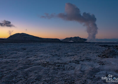 A panoramic view of a desolate landscape at dusk, featuring a rocky terrain with a texture resembling ash or gravel. In the background, rolling hills are silhouetted against a gradient sky transitioning from orange to deep blue. A plume of steam rises from a vent on the left side of the image, contributing to the atmospheric scene. The foreground includes a prominent, uneven surface with various rocky formations.