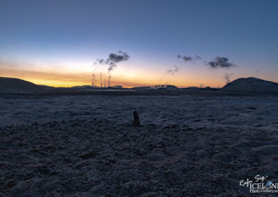 A vast barren landscape at dusk, featuring a textured ground with a rocky formation in the foreground. In the background, low hills are silhouetted against a gradient sky transitioning from deep blue to orange, with steam rising from geothermal features that create clouds above the horizon.