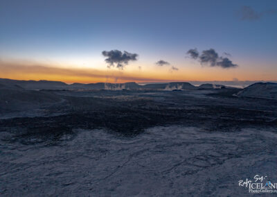 A vast, rugged volcanic landscape under a twilight sky. The ground is covered with dark, textured rock formations, while steam rises from several vents in the distance. The horizon showcases rolling hills silhouetted against a gradient of soft oranges and blues as the sun sets, casting a serene atmosphere over the unique terrain. Wispy clouds float above, adding to the ethereal quality of the scene.