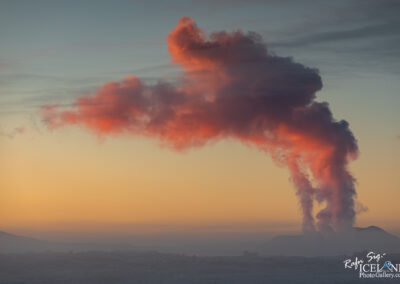 A picturesque scene at sunrise featuring soft pastel hues in the sky, with prominent clouds tinged in shades of pink and orange. In the foreground, wisps of steam rise from a geothermal vent against a backdrop of rugged hills and a serene horizon, creating a tranquil and atmospheric landscape.