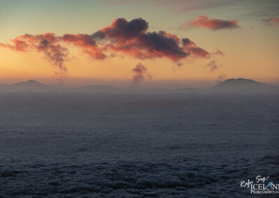 A serene landscape at sunset featuring a gradient sky with hues of orange and soft pink clouds. In the foreground, a textured, frost-covered terrain stretches out, while distant mountains rise gently against the horizon. Wisps of steam or vapor emerge from the landscape, adding a mystical quality to the scene.