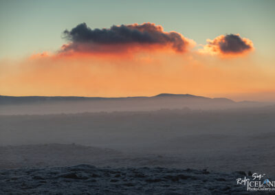 A scenic landscape featuring rolling hills and a distant horizon under a colorful sky at sunset. The sky displays soft gradients of orange, pink, and blue, with a few clouds softly illuminated by the setting sun. Below, a misty ground is visible, suggesting a serene, tranquil environment. The overall atmosphere conveys a sense of calm and natural beauty.