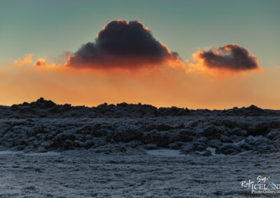 A landscape featuring a textured, rocky terrain covered in frost or snow, illuminated by a dramatic sunset. The sky transitions from soft blue to warm orange and deepening shadows, with a few clouds highlighted by the setting sun. The foreground displays rugged rocks, while the background showcases a picturesque horizon.