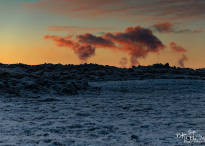 A striking landscape at sunset featuring a rocky terrain covered in frost, with steam rising into the sky. The horizon showcases vibrant hues of orange and purple, blending into a pale blue sky. The foreground presents a textured ground, while the background includes billowing clouds of steam, adding a mystical atmosphere to the scene.