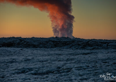 A vibrant sunset with shades of orange and blue casts a backdrop for an erupting volcano, sending a plume of smoke into the sky. The foreground features a textured, rocky landscape covered in frost, contrasting with the warm colors of the evening sky.