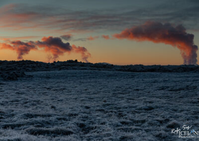 A serene landscape at dawn featuring a frosty terrain covered in white grass and small patches of rocky ground. In the background, two plumes of steam rise into the sky, blending with soft pink and orange clouds, creating a tranquil yet striking atmosphere against the lightening sky.