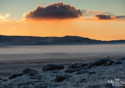 A beautiful landscape at dawn featuring rolling hills and distant mountains under a colorful sky. The horizon glows with shades of orange and light blue, while a thick mist blankets the ground. Frost-covered rocks are visible in the foreground, adding to the serene and chilly atmosphere.