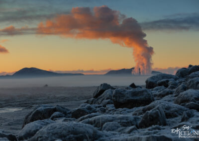 A panoramic view of a mountainous landscape at sunrise, with a prominent steam plume rising from a geothermal area in the background. The foreground features rugged, frost-covered rocks, while mist blankets the lower terrain, blending into the serene sky filled with pastel hues of orange and blue. The scene conveys a sense of tranquility and natural beauty.