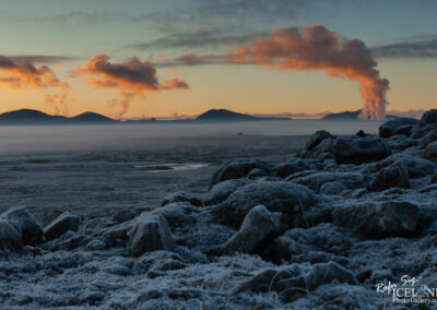 A breathtaking landscape showcasing a frozen terrain with scattered rocks in the foreground, leading to a flat, mist-covered expanse. In the background, a range of mountains is visible, partially shrouded in low-hanging fog. The sky is painted in warm hues of orange and blue as the sun sets, with wisps of steam rising from geothermal vents on the mountains, creating a striking contrast against the cool colors of the landscape.