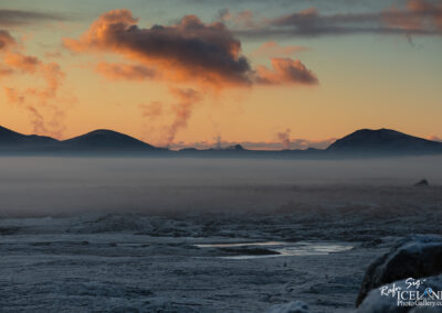 A tranquil landscape at sunset featuring rolling hills in the background, with soft clouds illuminated in warm tones. Mist rises from the ground, blending into the horizon where the hills meet the sky. The foreground showcases a textured, rocky terrain, partially covered in frost or snow.