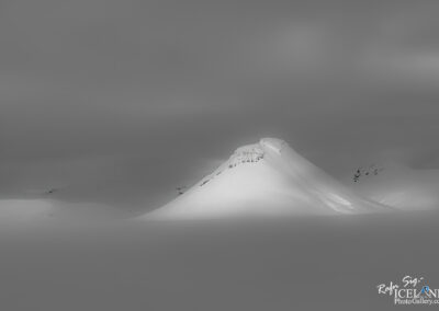 A snowy mountain rises above a flat, white landscape, under a predominantly gray sky. The mountain has a smooth, rounded peak with a slight crest, and its slopes are partially illuminated, creating a contrast against the surrounding monochromatic scenery. The atmosphere appears tranquil and desolate, emphasizing the vastness of the snow-covered expanse.