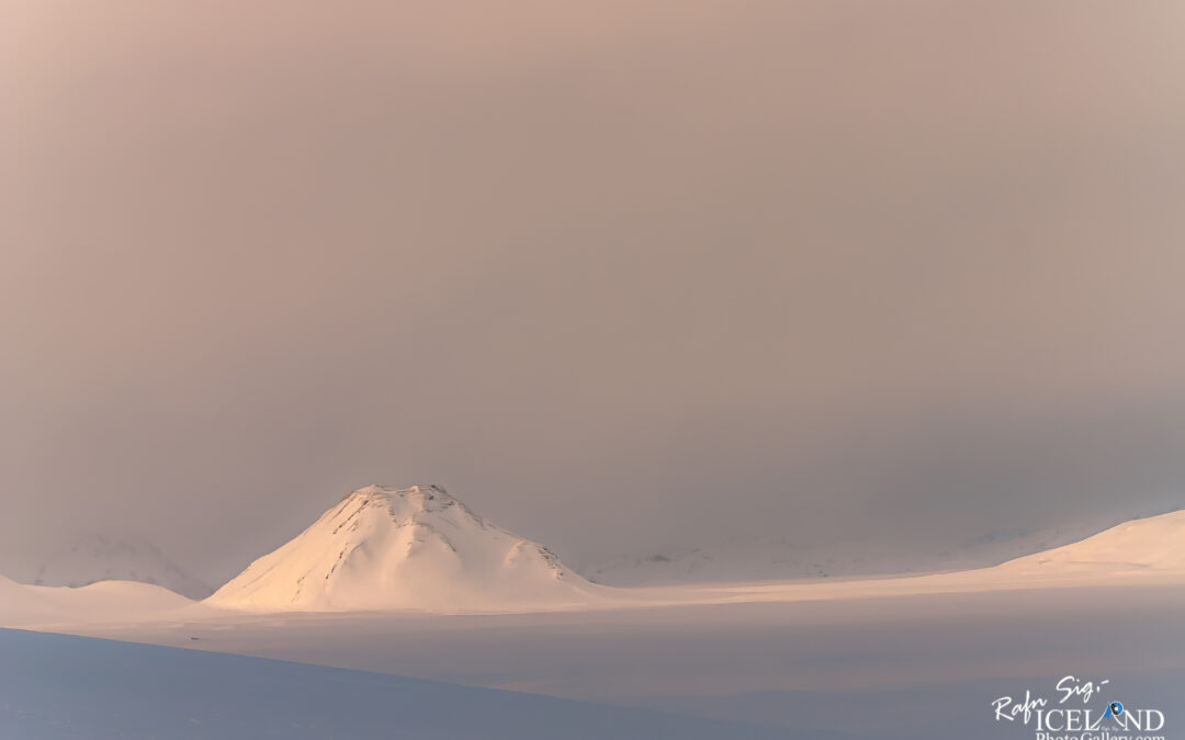 A serene winter landscape featuring snow-covered mountains shrouded in mist. The scene captures soft pastel tones in the sky, with the peaks faintly visible against a cloudy backdrop. The surface below is mostly flat and white, suggesting an expansive snowy area. The overall atmosphere is tranquil and ethereal, evoking a sense of remote beauty.
