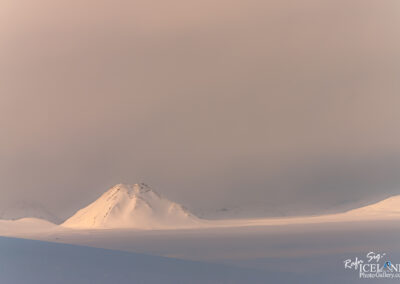 A serene winter landscape featuring snow-covered mountains shrouded in mist. The scene captures soft pastel tones in the sky, with the peaks faintly visible against a cloudy backdrop. The surface below is mostly flat and white, suggesting an expansive snowy area. The overall atmosphere is tranquil and ethereal, evoking a sense of remote beauty.