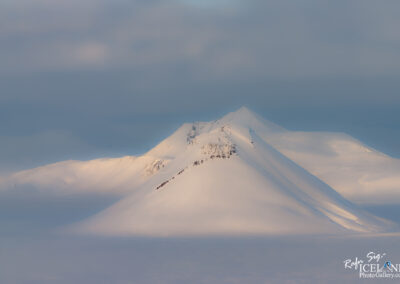 A serene landscape featuring a snow-covered mountain peak, partially illuminated by soft light, with a cloudy sky in the background. The scene conveys a tranquil and expansive natural beauty, highlighting the contours of the slopes and the vastness of the surrounding area blanketed in snow.