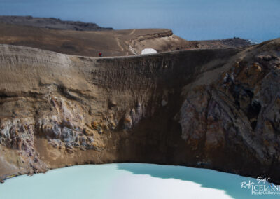 The image showcases a striking volcanic landscape featuring a large, crater-like structure with steep, textured sides. The interior of the crater contains a vibrant turquoise lake, its surface gently rippling. A few individuals are seen walking along the edge of the crater, providing a sense of scale to the vastness of the scene. The terrain surrounding the crater is barren, with earthy tones of brown and hints of mineral colors on the rocky walls. In the background, a serene body of water can be glimpsed, contrasting with the rugged landscape. The sky above is clear, completing this dramatic natural setting.