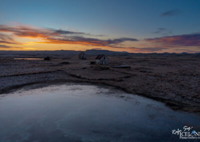 A serene landscape at sunset, showcasing a vast, open terrain characterized by dry grass and rocky patches. In the foreground, a calm body of water reflects the soft hues of the sky, which transitions from deep blue to vibrant orange and pink as the sun sets. To the right, two small, weathered houses with sloping roofs stand in isolation, their white walls contrasting with the natural surroundings. The distant horizon is lined with gentle hills, partially shrouded in a soft mist, while wispy clouds drift across the sky, adding texture to the tranquil scene. The overall atmosphere is one of peaceful solitude in a remote, rugged environment.