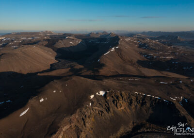 A panoramic view of a rugged mountainous landscape under a clear blue sky. The terrain features a mix of dark earth tones, with gently rolling hills and sharp ridges casting long shadows across the ground. Scattered patches of white snow cling to some of the slopes, contrasting with the rocky surfaces. The landscape appears vast and somewhat barren, with hints of texture created by the undulating hills. In the distance, other mountain peaks rise, adding depth to the scene. Overall, it conveys a sense of wild natural beauty.