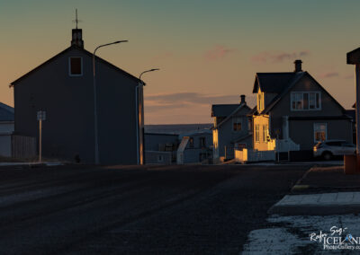 A quiet street scene at sunset, featuring a row of houses with simple architecture. On the left, a dark gray building stands with a pointed roof and a small antenna. Nearby, a white-flanked pathway leads to a pale gray house with a peaked roof and multiple windows. The houses are softly illuminated by the warm light of the setting sun, creating long shadows on the pavement. A few street lamps are positioned along the road, and a silver car is parked beside one of the houses. In the background, there are hints of the open sky and possibly water, with distant, muted hues blending into one another.