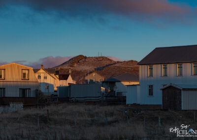 A scenic view of a small town with several houses, mostly white and light-colored, clustered together. The buildings have a simple, contemporary design with some featuring metal siding and large windows. In the background, there’s a rocky hill topped with antennas, partially dusted with snow, against a backdrop of soft pink and blue clouds. The foreground has tall, dry grass and some fences, adding to the quiet, rural atmosphere of the setting. The lighting suggests either early morning or late afternoon, casting warm tones on the structures.