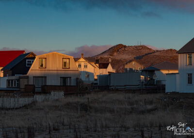 The image shows a peaceful scene of a small town at dusk. Various buildings stand close together, some with sloped roofs painted in colors like red and grey, while others have a lighter, metallic appearance. The sunlight casts warm tones on the sides of the structures, highlighting their textures. In the background, a mountainside can be seen, with a few patches of snow visible and a communications tower near the summit. The foreground features sparse grass and a white picket fence separating the houses from the open land, creating a cozy, inviting atmosphere. The sky is a blend of blue and soft pink hues, suggesting the end of the day.