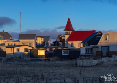 The image showcases a cluster of buildings at sunset, highlighting a mix of architectural styles. In the foreground, several smaller, dark-painted houses and a larger light-colored building catch the warm glow of the setting sun. A prominent church with a striking red roof and a tall steeple stands out against the evening sky. The background is filled with various other houses, some with pitched roofs and others flat, creating a quaint, community feel. Above, a tall communications tower rises, blending into the blue and cloudy sky. The scene conveys a sense of tranquility and charm, typical of a small town.
