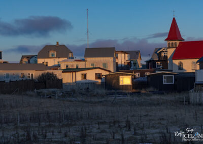 The image captures a quiet neighborhood scene during twilight. Various houses, primarily made of wood, are visible, each featuring distinct architecture with slanted roofs and a mix of colors, including white and muted tones. A prominent red-roofed church stands out among the houses, topped with a small cross. In the foreground, there is sparse vegetation and a few wooden fences, creating a sense of openness. The setting sun casts soft shadows and highlights the textures of the buildings, while a light breeze stirs the atmosphere. The overall mood is serene and reflective, evoking a peaceful, small-town ambiance.