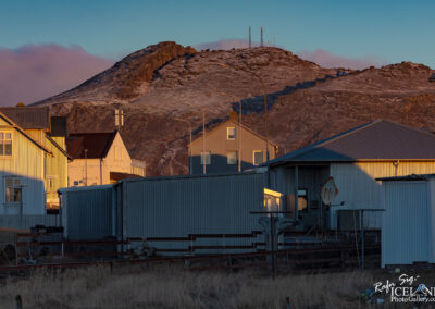 The image depicts a landscape with a mix of residential buildings and a mountainous backdrop. In the foreground, several houses with varying architectural styles and colors, predominantly white and beige, are visible. Some buildings are partially shaded, while others catch the warm hues of sunlight, enhancing their details. In the background, a rugged mountain stands tall, its surface showing signs of snow and rocky terrain, with a few telecommunications towers perched at the summit. The sky above transitions from a soft blue to warm tones, indicating either sunrise or sunset, creating a serene atmosphere complemented by sparse vegetation in the vicinity.
