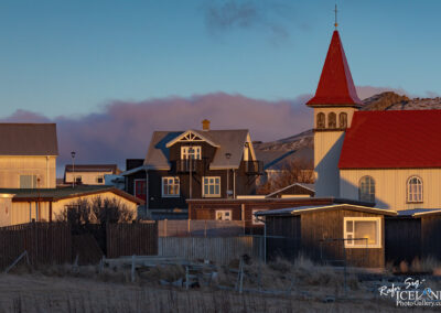 The image captures a quaint village scene during the golden hour. In the foreground, there are various houses with distinct styles, including one with a modern dark wood exterior and large windows, and another with a bright red roof. A few small trees and patches of grass are scattered among the homes, contributing to the natural feel of the setting. To the right, there is a tall white church with a striking red spire topped by a small cross, standing out against the backdrop of soft, colorful clouds in the sky. The overall ambiance is serene, evoking a peaceful rural atmosphere.