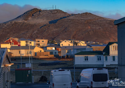 A scenic view of a small town with colorful houses nestled at the foot of a rocky, snow-dusted mountain. The homes, primarily white with red and green accents, are arranged closely together and bathed in warm golden light, suggesting early morning or late afternoon. There are several light poles and a green-roofed structure, possibly a shed or a small building, visible in the foreground. In the background, the mountain rises sharply, covered in patches of snow and rocky terrain, with some communication towers at its peak. The atmosphere is serene and picturesque, embodying a sense of quiet rural life.